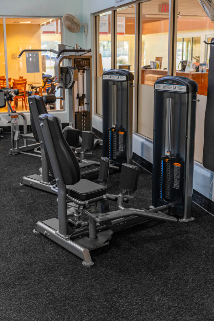 Two black gym machines for leg exercises stand side by side on a carpeted floor in a fitness center, with large windows showing an office area in the background.