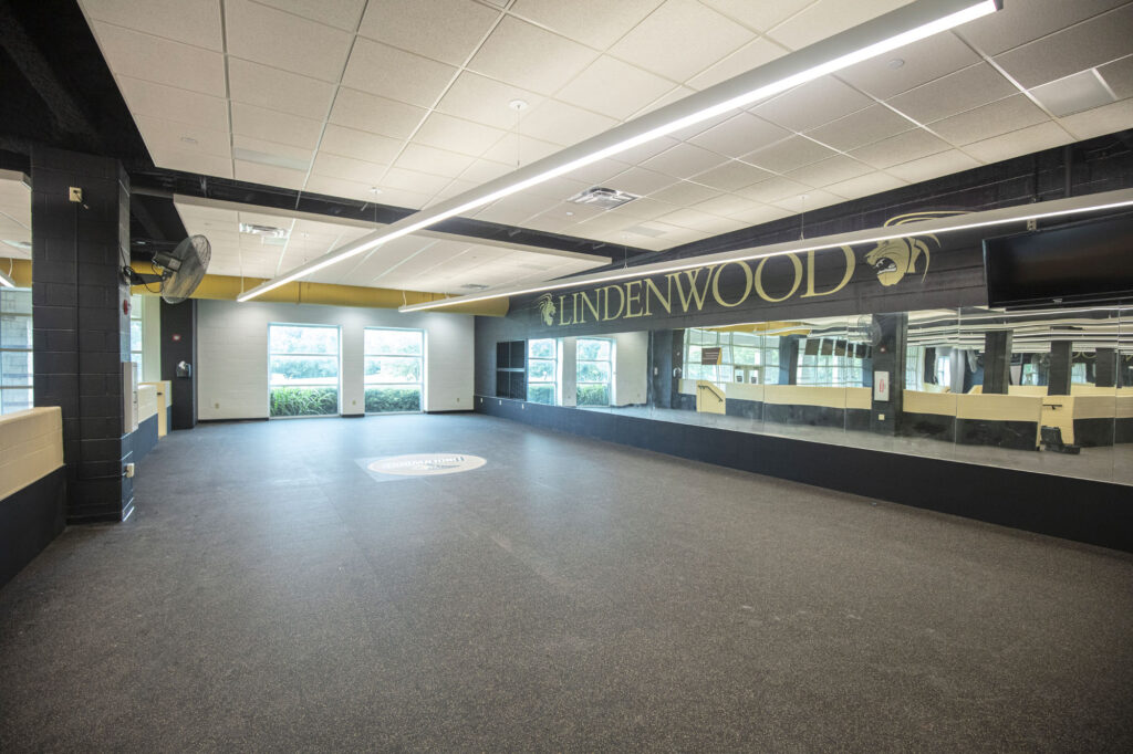 Spacious, empty exercise room with large mirrors, black and gold walls, Lindenwood and lion logo on the wall, windows letting in natural light, and a TV mounted in the corner.