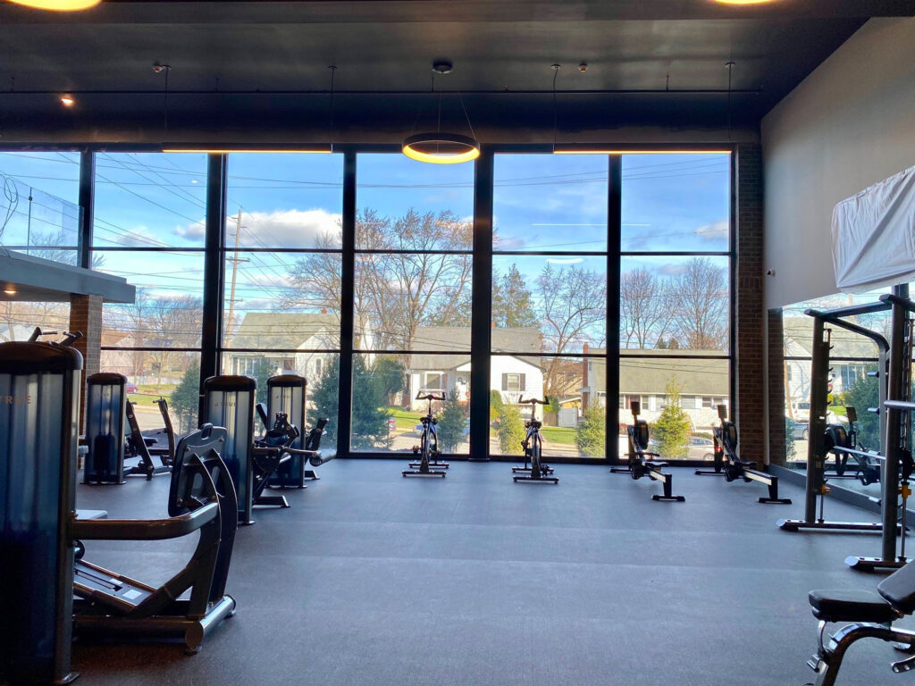 A modern gym with large floor-to-ceiling windows, exercise bikes, rowing machines, and various fitness equipment. Outside, trees and houses are visible under a blue sky with scattered clouds.