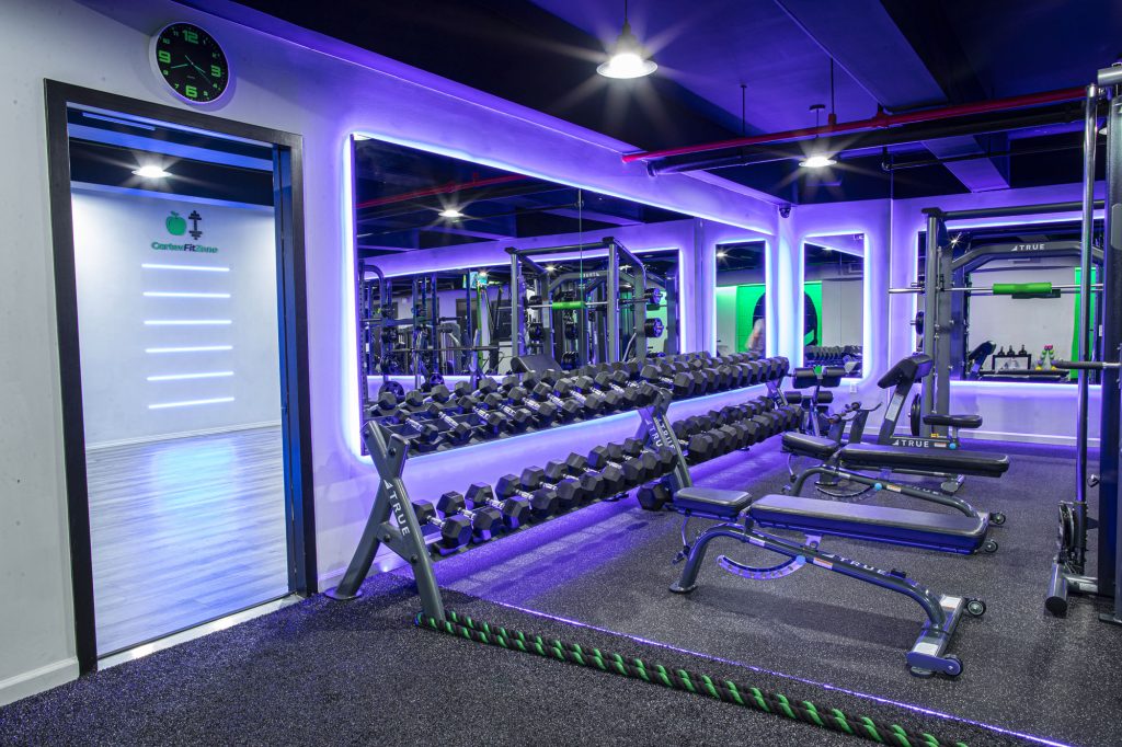 A modern gym with purple LED lighting, featuring mirrored walls, racks of dumbbells, benches, cardio machines, and a rope. A clock and an entrance to another room are visible on the left side.