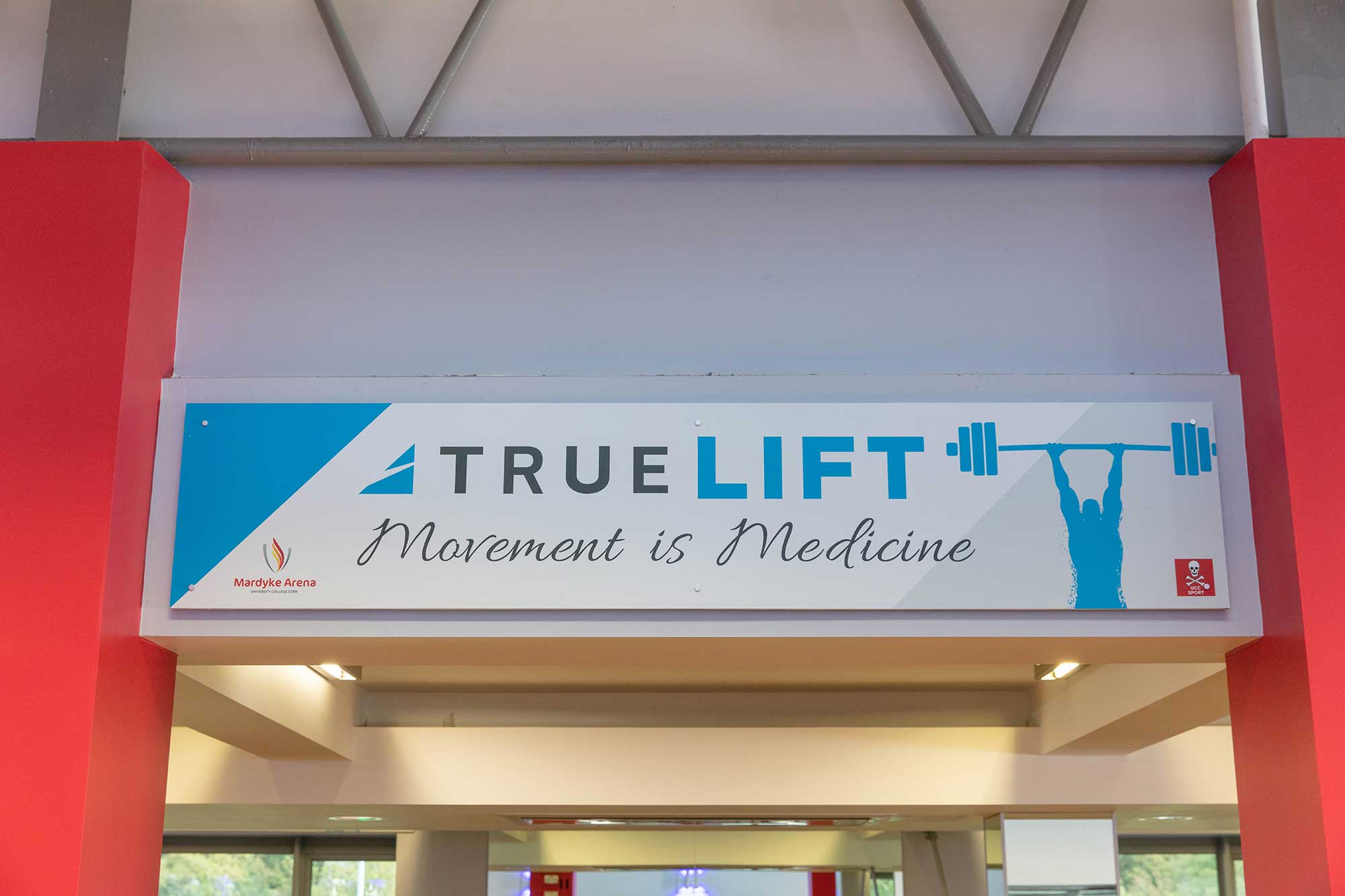 A sign above a gym entrance reads TRUE LIFT Movement is Medicine with a silhouette of a person lifting a barbell. The sign is bordered by red pillars and features gym-related branding.