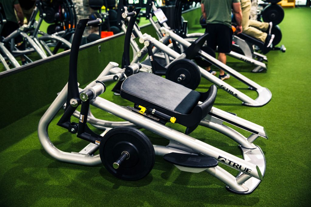 A seated exercise machine with weights and handles is positioned on artificial grass in a gym, with other fitness equipment and people in the background.