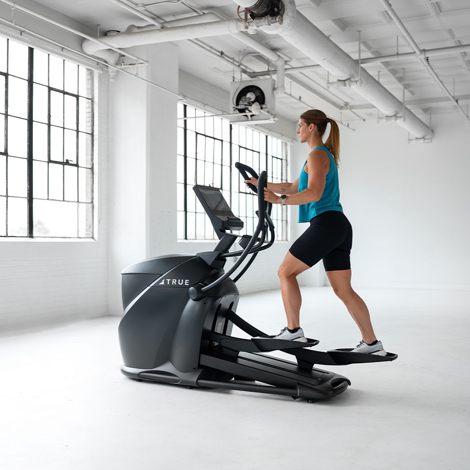 A woman in athletic wear exercises on an elliptical machine in a bright, spacious gym with large windows and white walls.