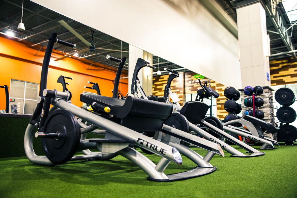Row of modern gym exercise machines on green turf, with an orange wall, stacked weights, and medicine balls visible in a spacious, well-lit fitness center.