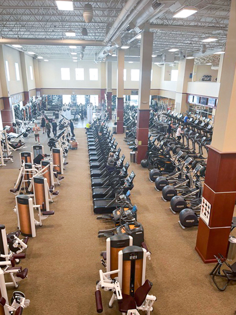 Large, spacious gym with rows of treadmills, ellipticals, stationary bikes, and various strength training machines. A few people are using the equipment. High ceilings and large windows let in natural light.