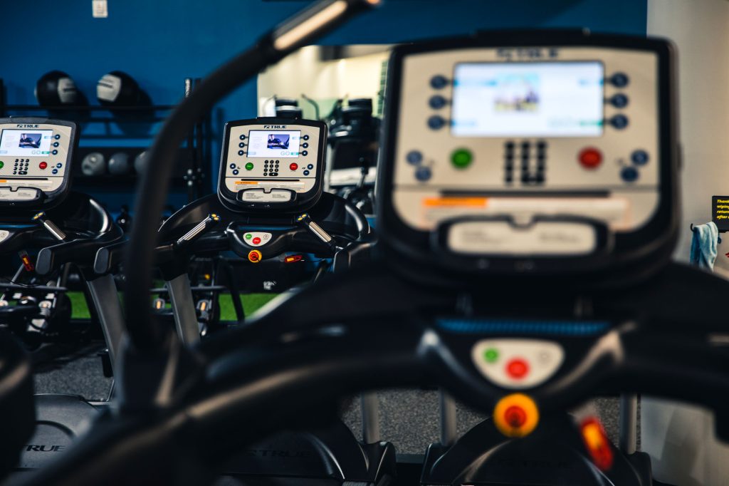 Close-up of three treadmill control panels in a gym, with weights and workout equipment visible in the background. The focus is on the front treadmill’s console, displaying various buttons and screens.