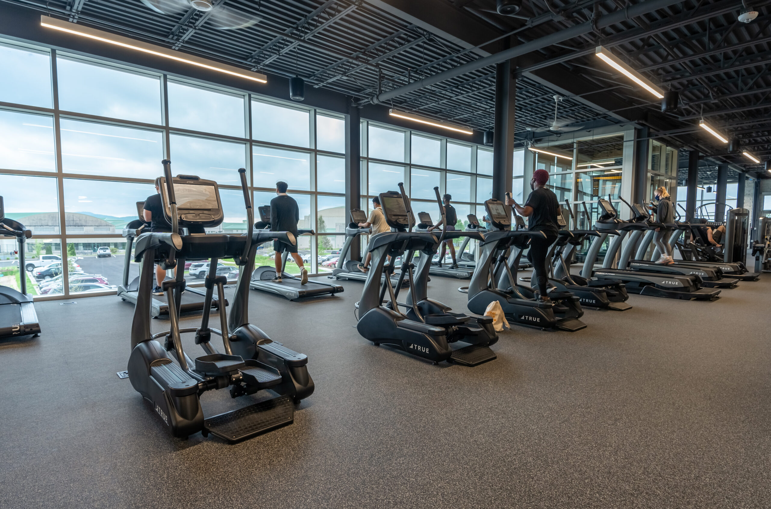 A row of people exercise on treadmills and elliptical machines in a modern gym with large windows, letting in natural light and offering views of the outdoors.