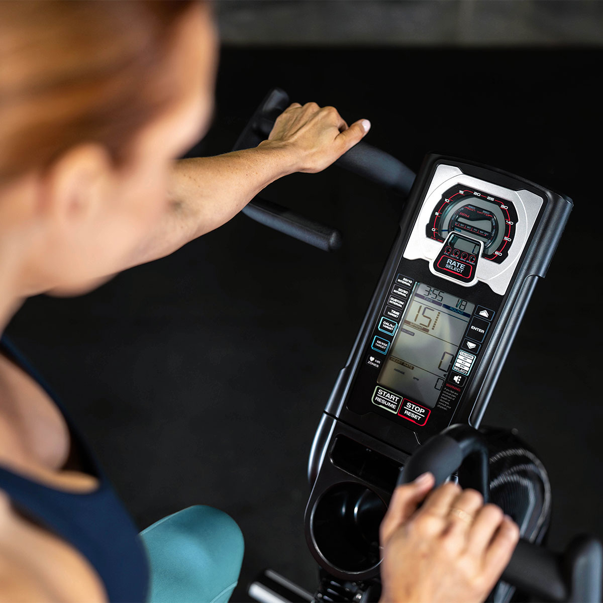 A person uses a stationary exercise bike, gripping the handlebars. The bike’s digital display shows various workout metrics, including time, speed, and distance. The background is blurred.