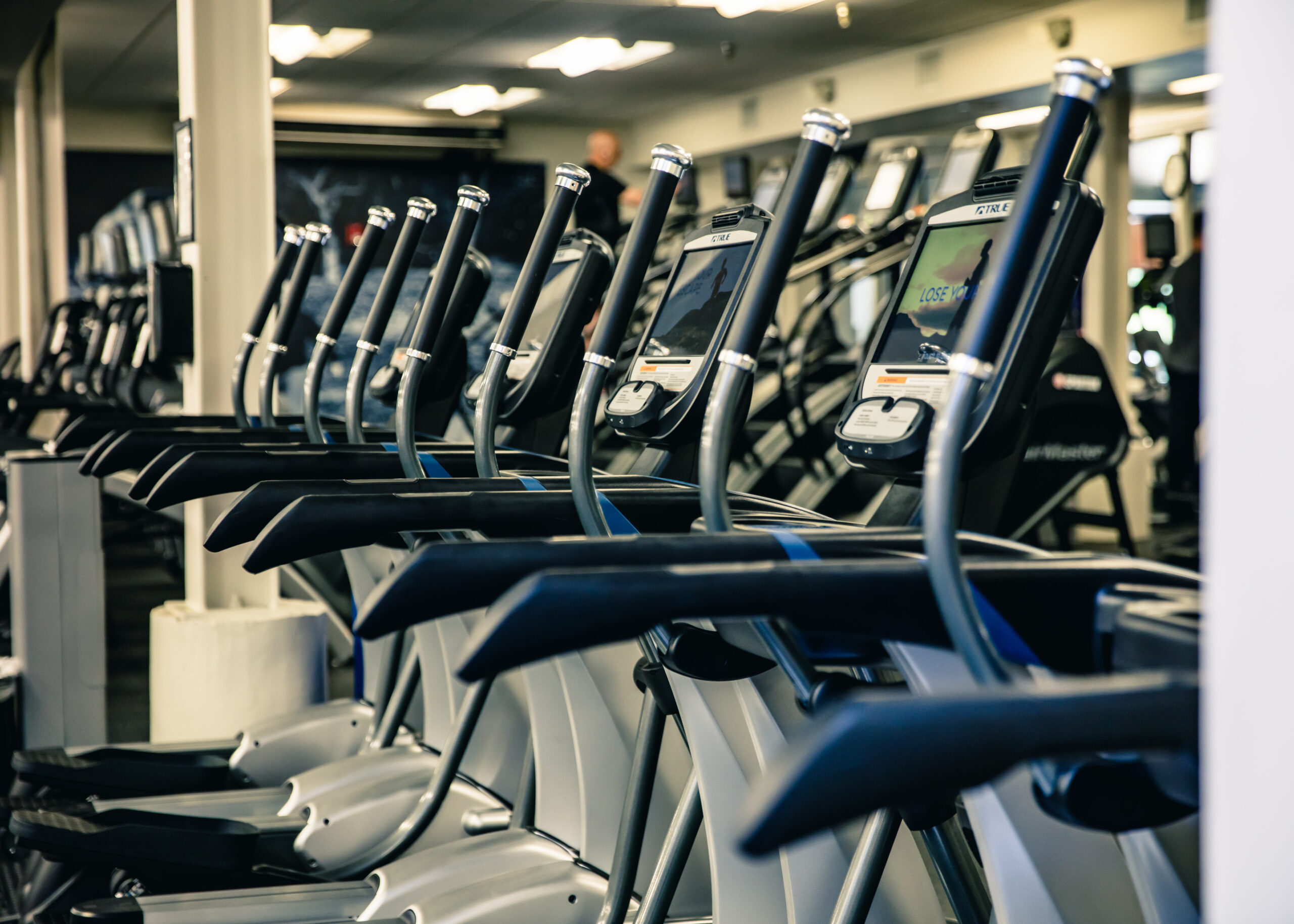 A row of elliptical machines lined up in a brightly lit gym, with screens and handles visible in the foreground. The gym appears clean and empty, with a few people blurred in the background.