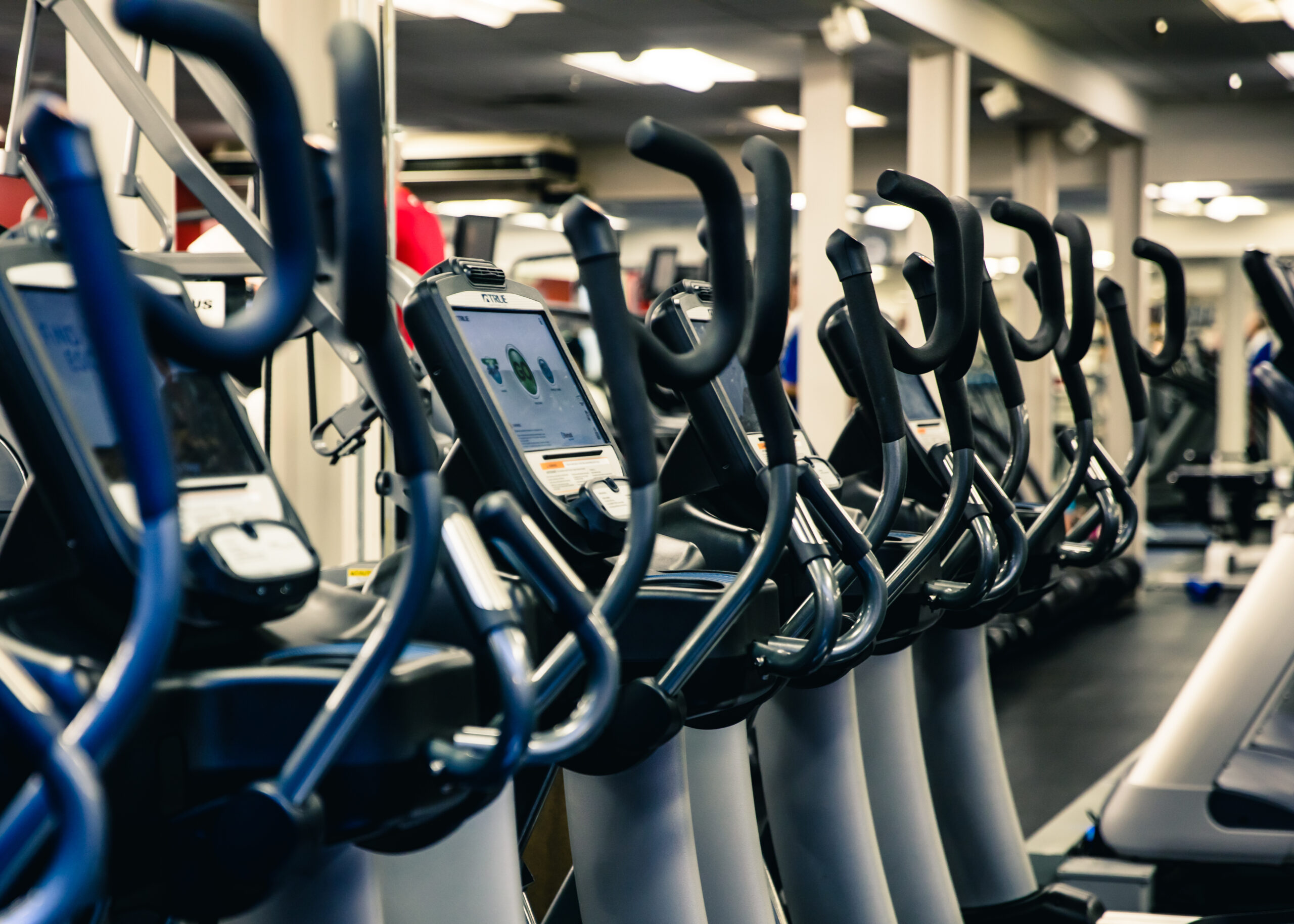 Row of elliptical machines in a gym, lined up closely together with digital screens and handlebars in focus, under bright ceiling lights and surrounded by mirrors and equipment.