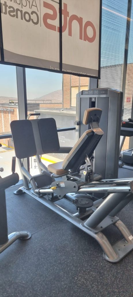 A seated leg press machine in a gym, positioned near large windows with sunlight streaming in. The gym has black flooring and visible exercise equipment in the background.