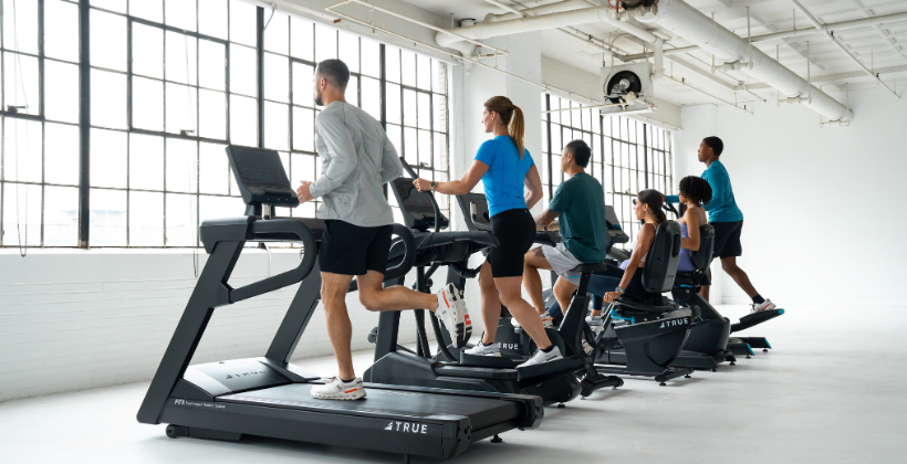 A group of people exercise inside a bright gym, using treadmills and stationary bikes arranged in a row near large windows that let in natural light.