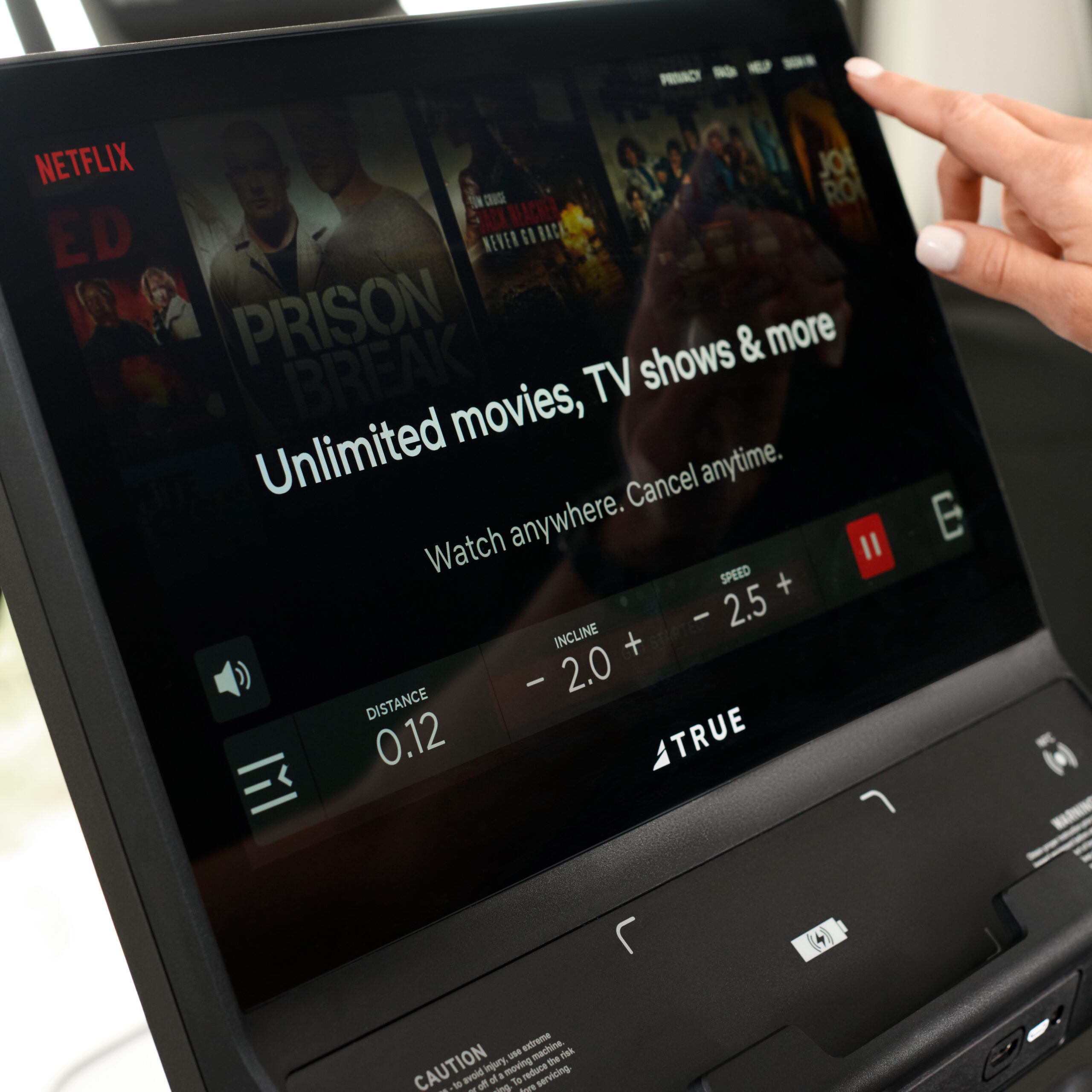A close-up of a treadmill screen displaying Netflix with the message “Unlimited movies, TV shows & more” as a person’s hand touches the screen. Various show thumbnails are visible in the background.
