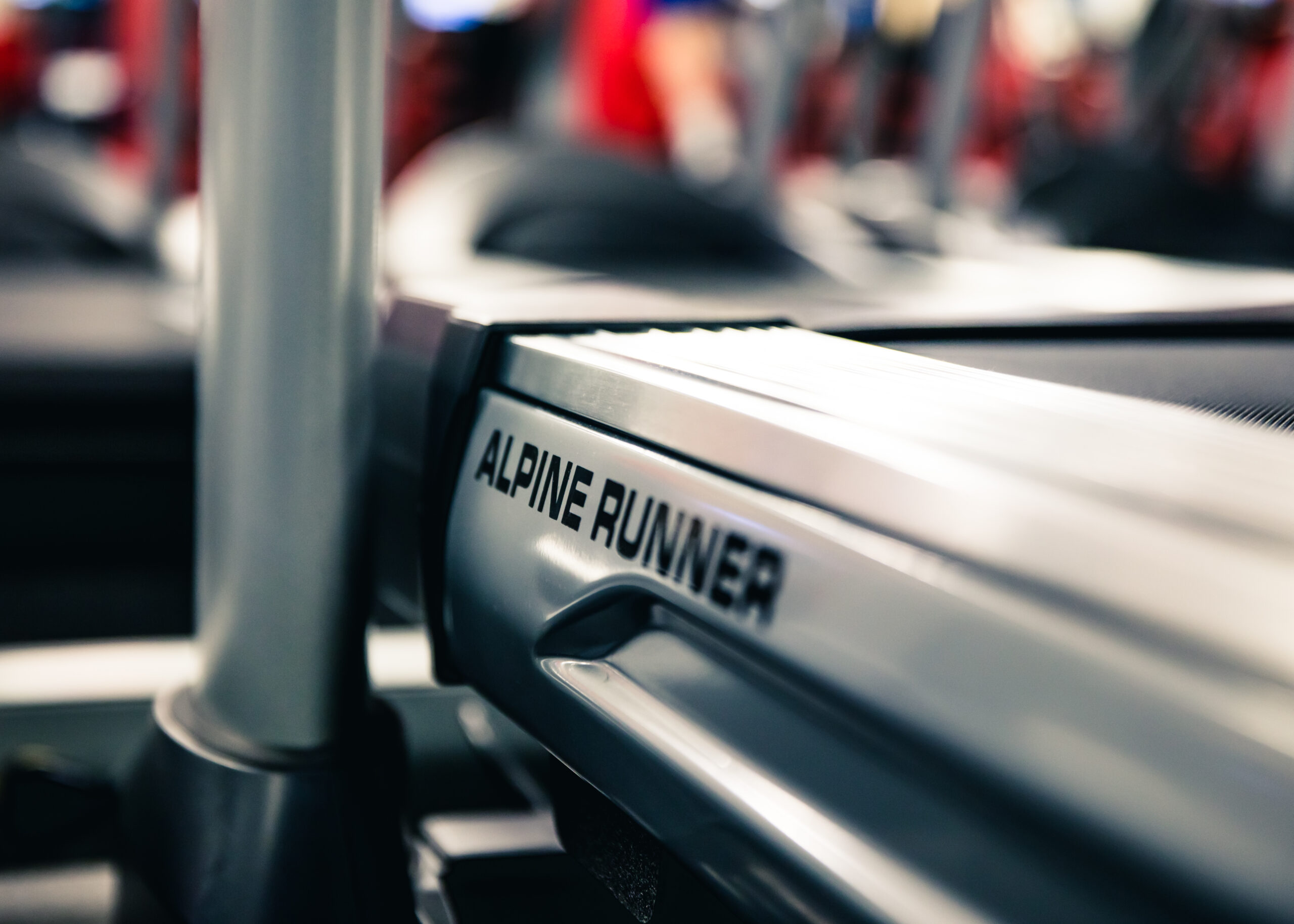 Close-up of a treadmill with the brand name Alpine Runner visible on its side; the background is blurred with hints of a gym environment.