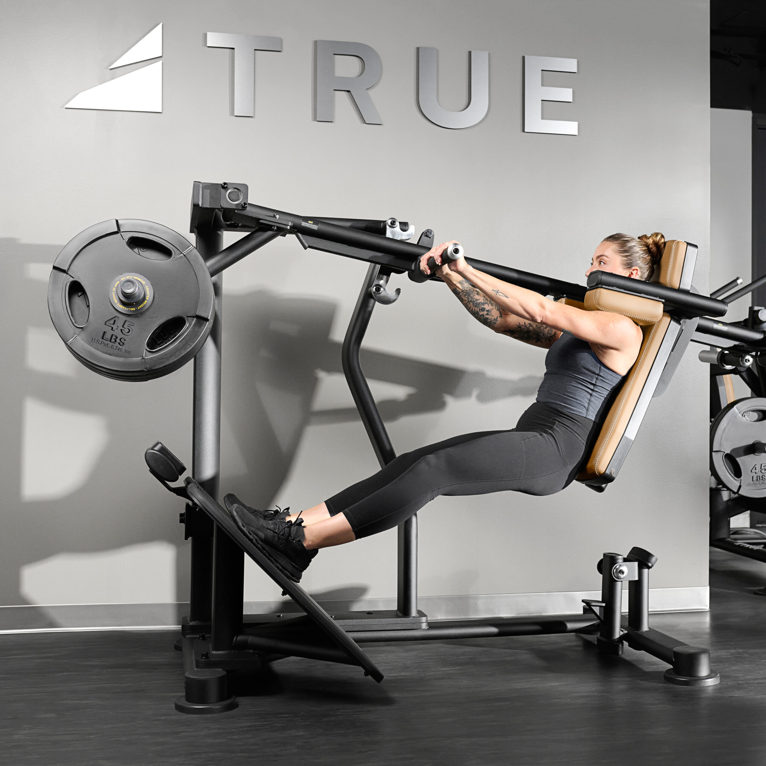 A woman in athletic wear uses a leg press machine in a gym, pressing a weighted platform with her legs. The wall behind her features the word TRUE in large letters.