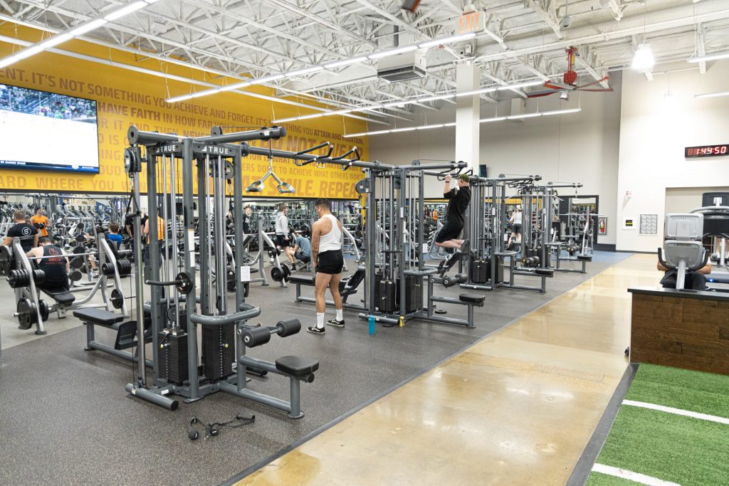 A spacious gym with people working out on various strength training machines. The area is well-lit, with motivational quotes on the yellow wall, and exercise equipment is neatly arranged on rubber flooring.