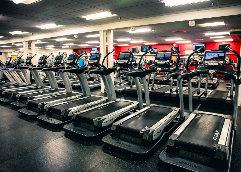 A row of empty treadmills is lined up in a brightly lit gym with red accent walls, black flooring, and several TVs mounted on the walls in the background.
