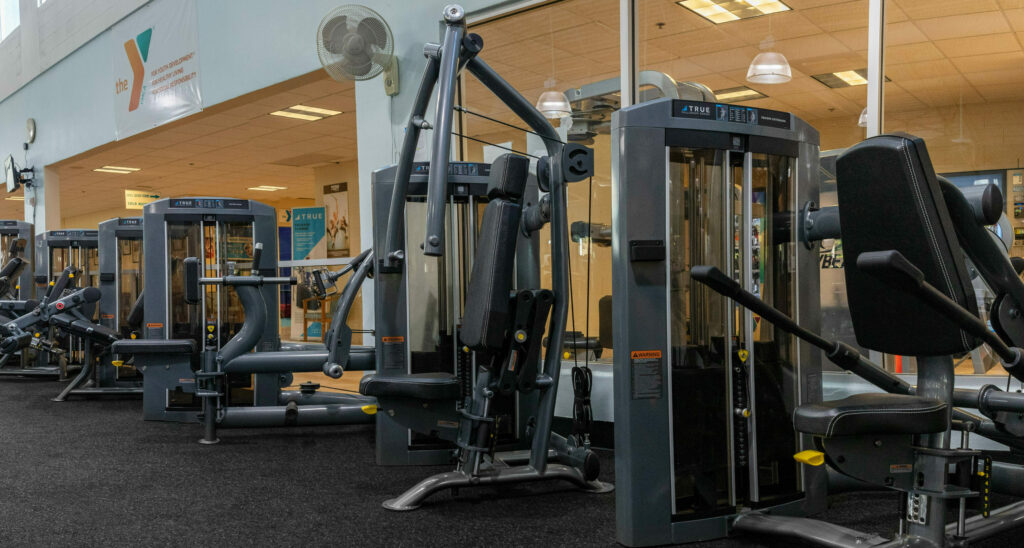 A row of strength training machines lines a gym with large windows, mirrors, and bright lighting. The machines are black and gray, and the gym appears clean and organized.