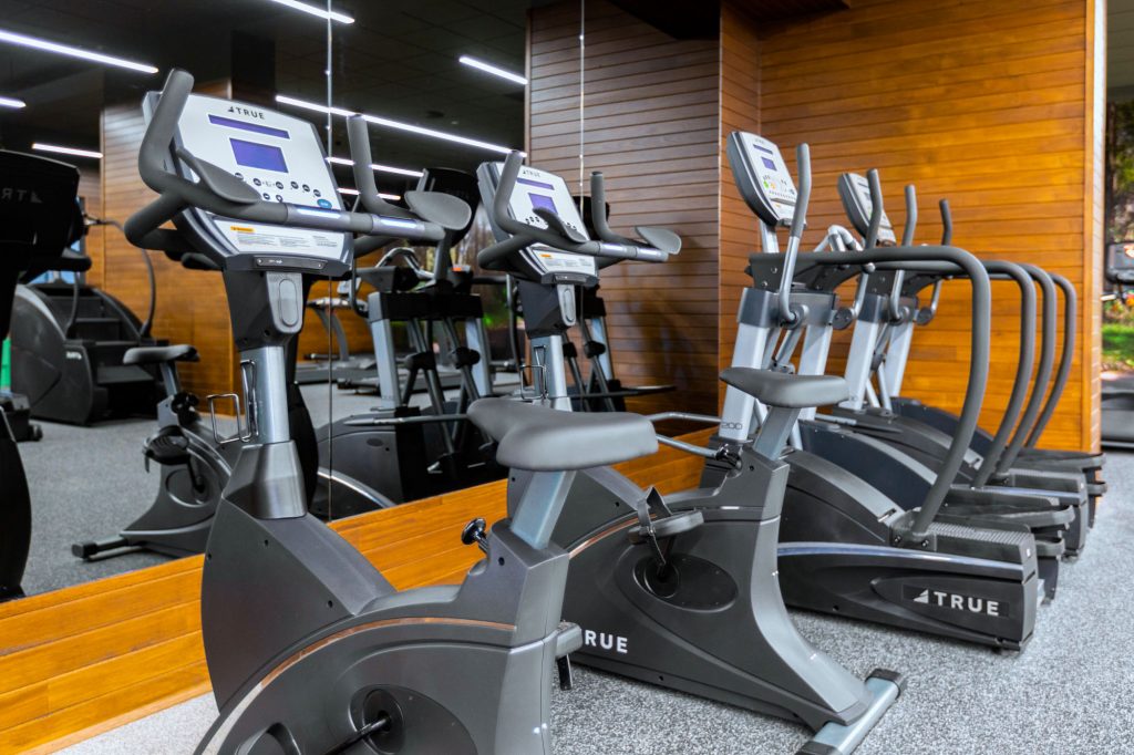Three stationary exercise bikes and two elliptical machines are lined up in a modern gym, with wooden wall paneling and large mirrors in the background.