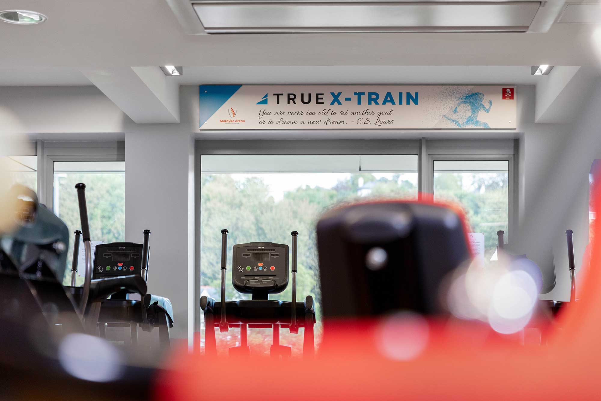 A gym interior with cardio machines facing large windows. Above the windows is a sign that reads TRUE X-TRAIN with a motivational quote and a blue graphic of a running figure.
