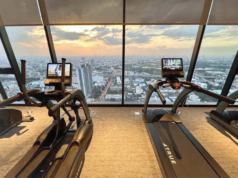 A modern gym with treadmills and ellipticals faces large floor-to-ceiling windows, offering a panoramic city skyline view at sunset, with buildings and glowing clouds in the background.