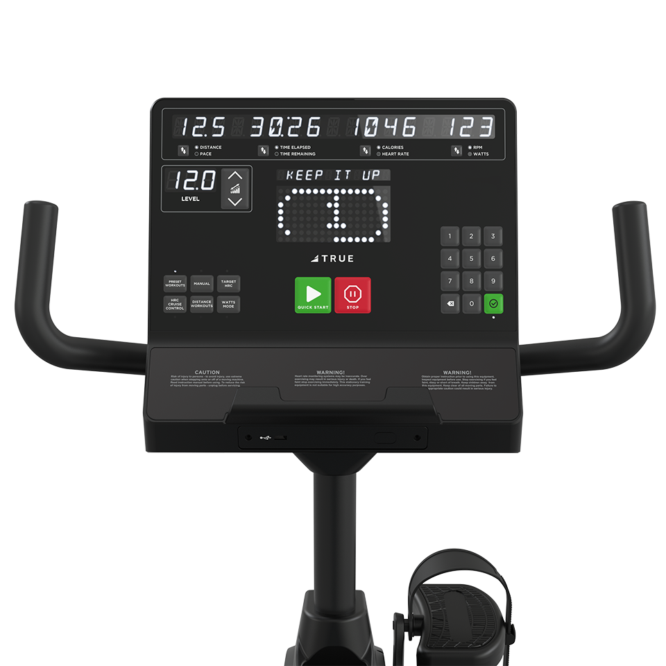 Close-up view of an exercise bike console displaying workout metrics, a message reading KEEP IT UP, various buttons, and handlebars on both sides. A foot pedal is partially visible at the bottom.