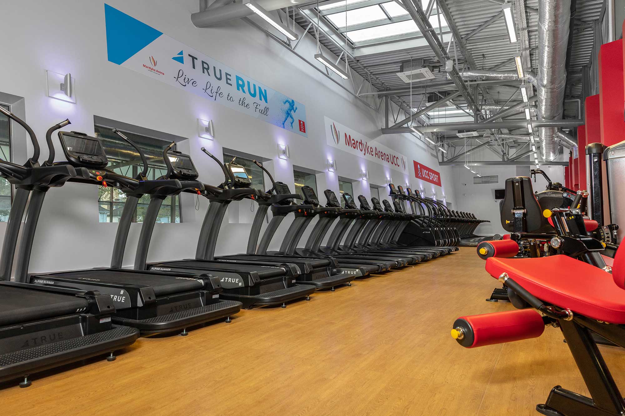 A modern gym with a row of treadmills along one wall, motivational signs above them, and strength training equipment with red padding in the foreground on a clean wooden floor.