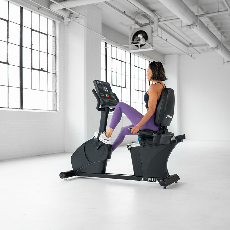 A woman in athletic wear uses a stationary recumbent exercise bike in a bright, spacious gym with large windows and white walls.