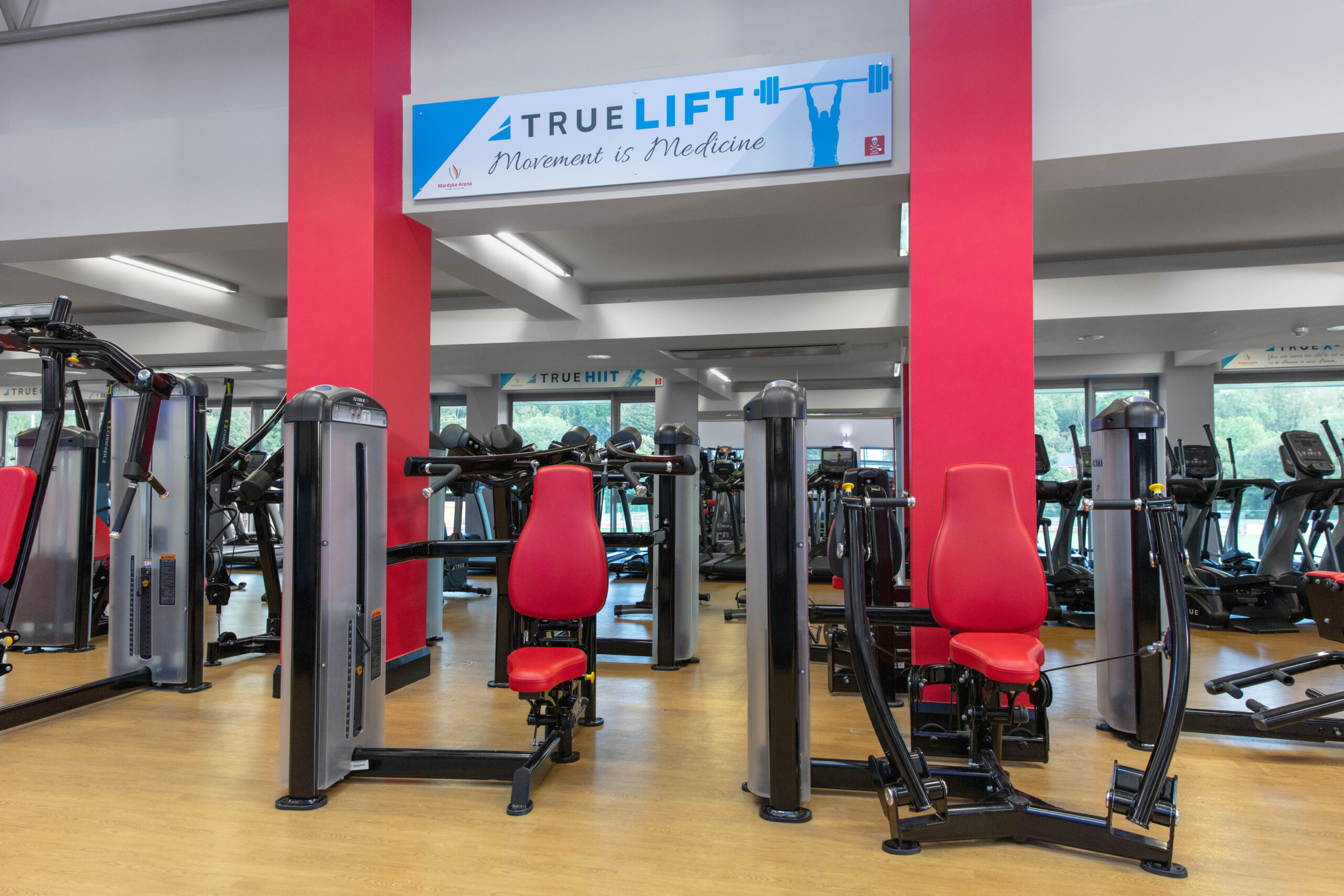 A brightly lit gym with red and gray exercise machines, wooden floors, and red columns. A sign above reads TRUELIFT Movement is Medicine. Large windows let in natural light.