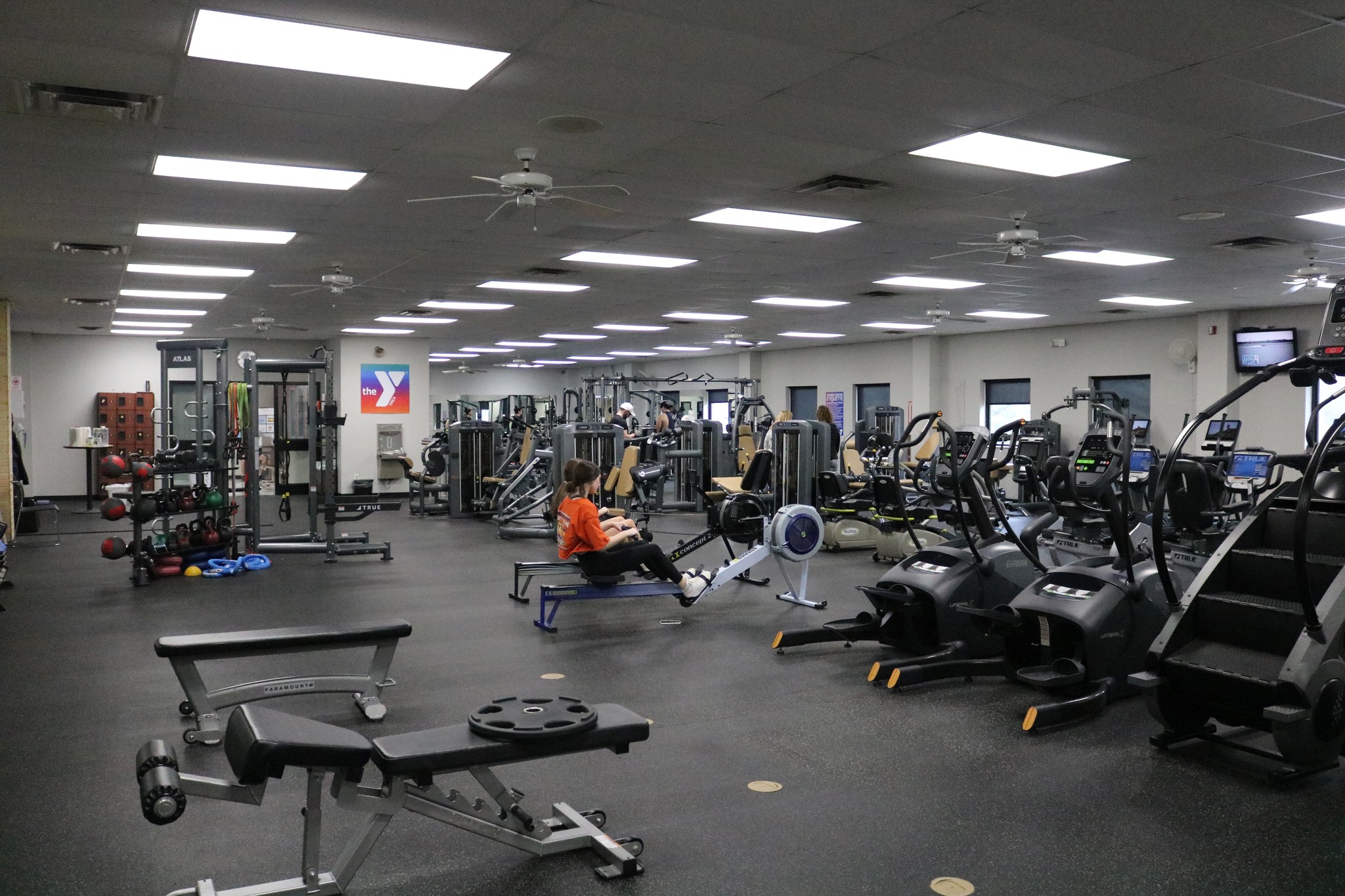 A spacious gym with various exercise machines, weights, and benches. One person in an orange shirt is using a rowing machine, while others use equipment in the background. The gym is well-lit with ceiling lights.
