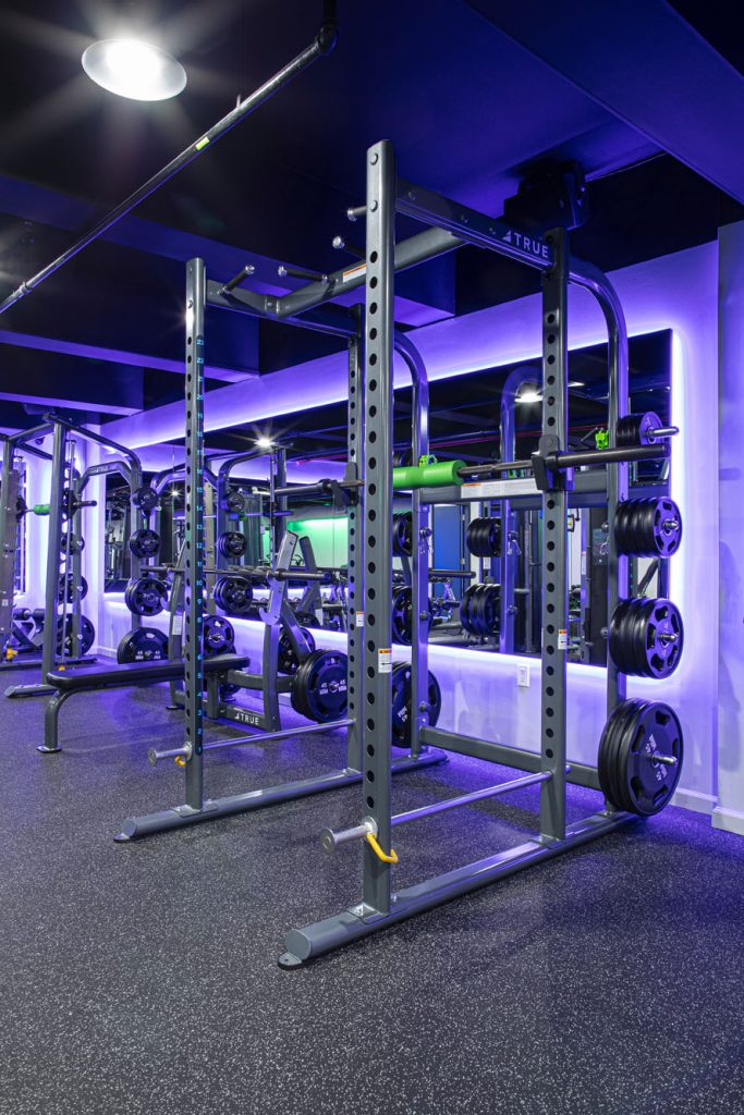 A modern gym with squat racks, barbells loaded with weight plates, and mirrors lining the walls, illuminated by blue and purple accent lighting. The floor has black rubber gym flooring.