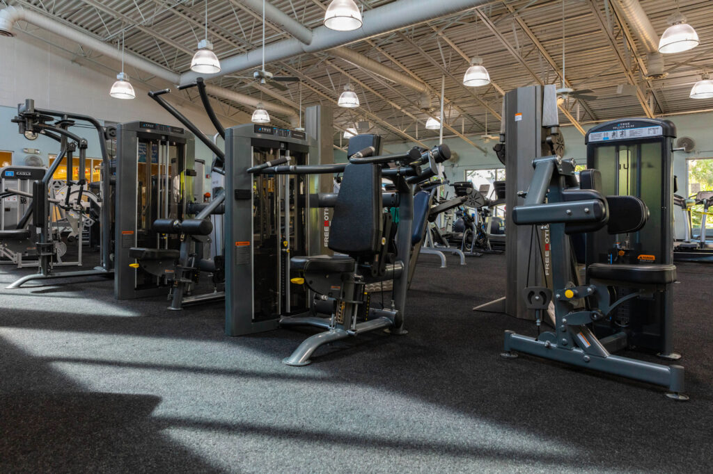 A spacious gym interior with several strength training machines neatly arranged on dark carpet flooring, surrounded by bright lighting and large windows letting in natural light.
