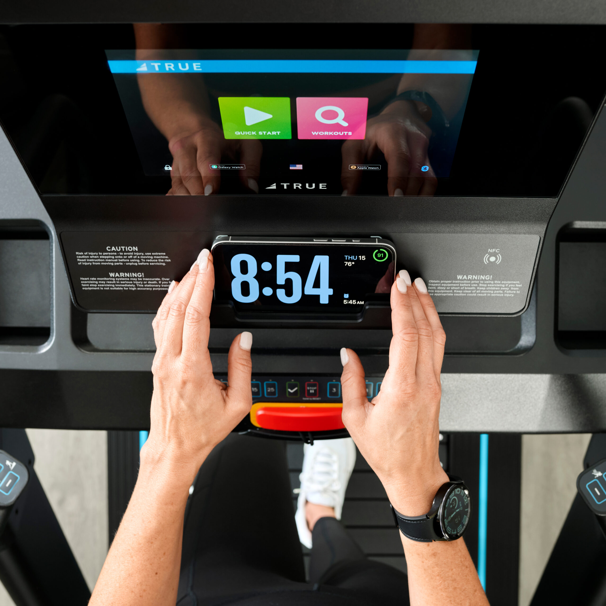 A person sets a smartphone with a large digital clock display on a treadmill console, which also shows touchscreen workout options. The person wears a black watch and holds the phone with both hands.