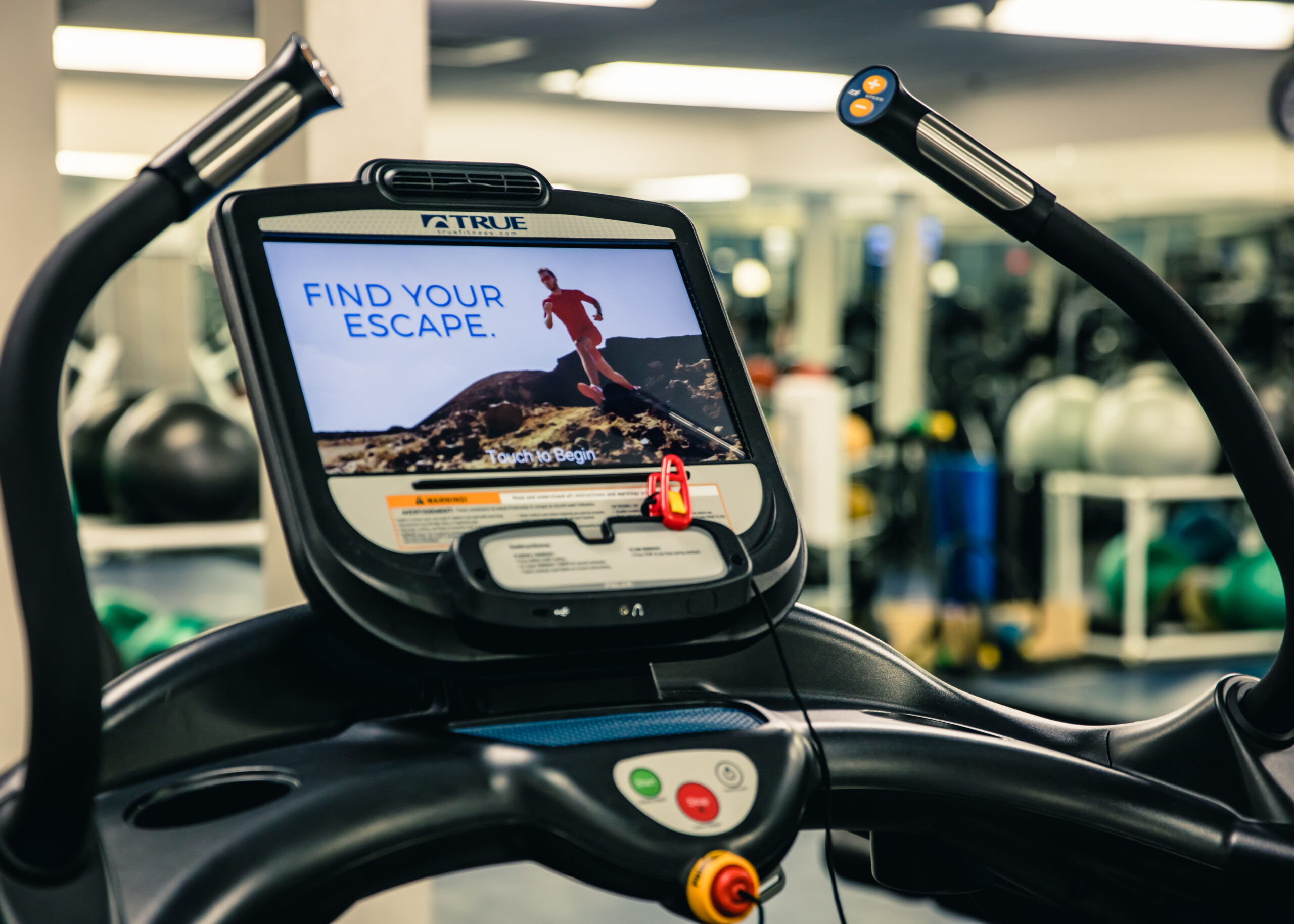 Close-up of a treadmill display screen at a gym, showing an advertisement with a person running outdoors and the words “Find Your Escape.” Exercise balls and gym equipment are visible in the blurred background.
