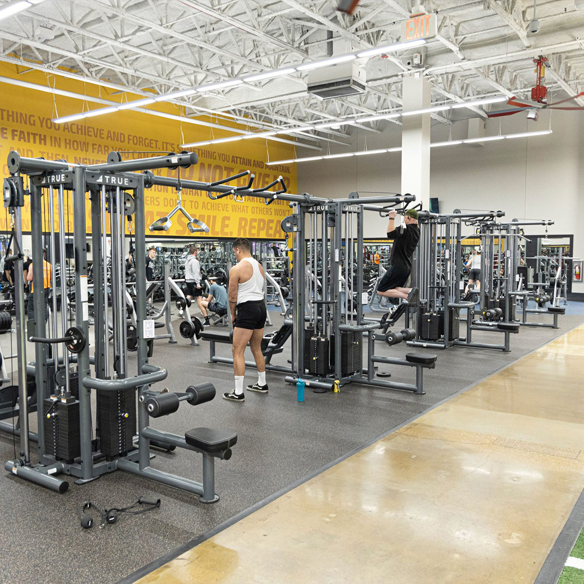 A spacious gym with several people exercising on weight machines and pull-up bars. The walls have motivational quotes in large yellow text. The gym has bright lighting and rubber flooring.