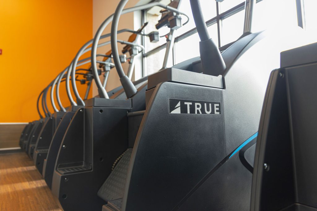A row of black stair stepper exercise machines is lined up in a gym with an orange accent wall and wood flooring; sunlight streams in through a window.
