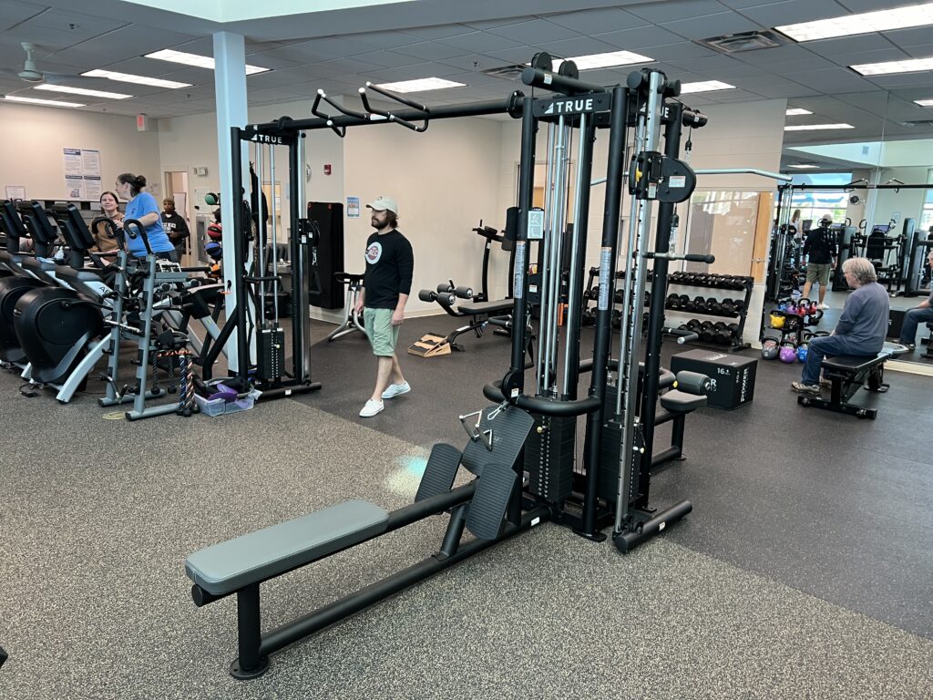 A spacious gym with various exercise machines, free weights, and people working out. One person adjusts equipment while others use cardio machines and weight benches in a bright, well-lit room.