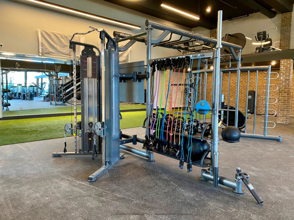 A gym with various exercise equipment, including a cable machine, colorful resistance bands, medicine balls, and turf in the background, all in a spacious, well-lit workout area with mirrored and brick walls.