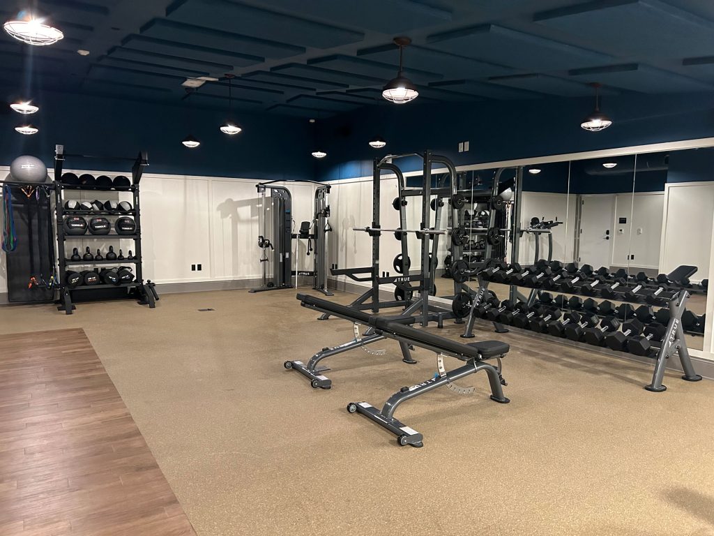 Modern gym with free weights, kettlebells, benches, squat rack, resistance bands, and exercise balls. A large mirror spans one wall; the floor is tan rubber and wood, with blue ceiling and bright overhead lights.