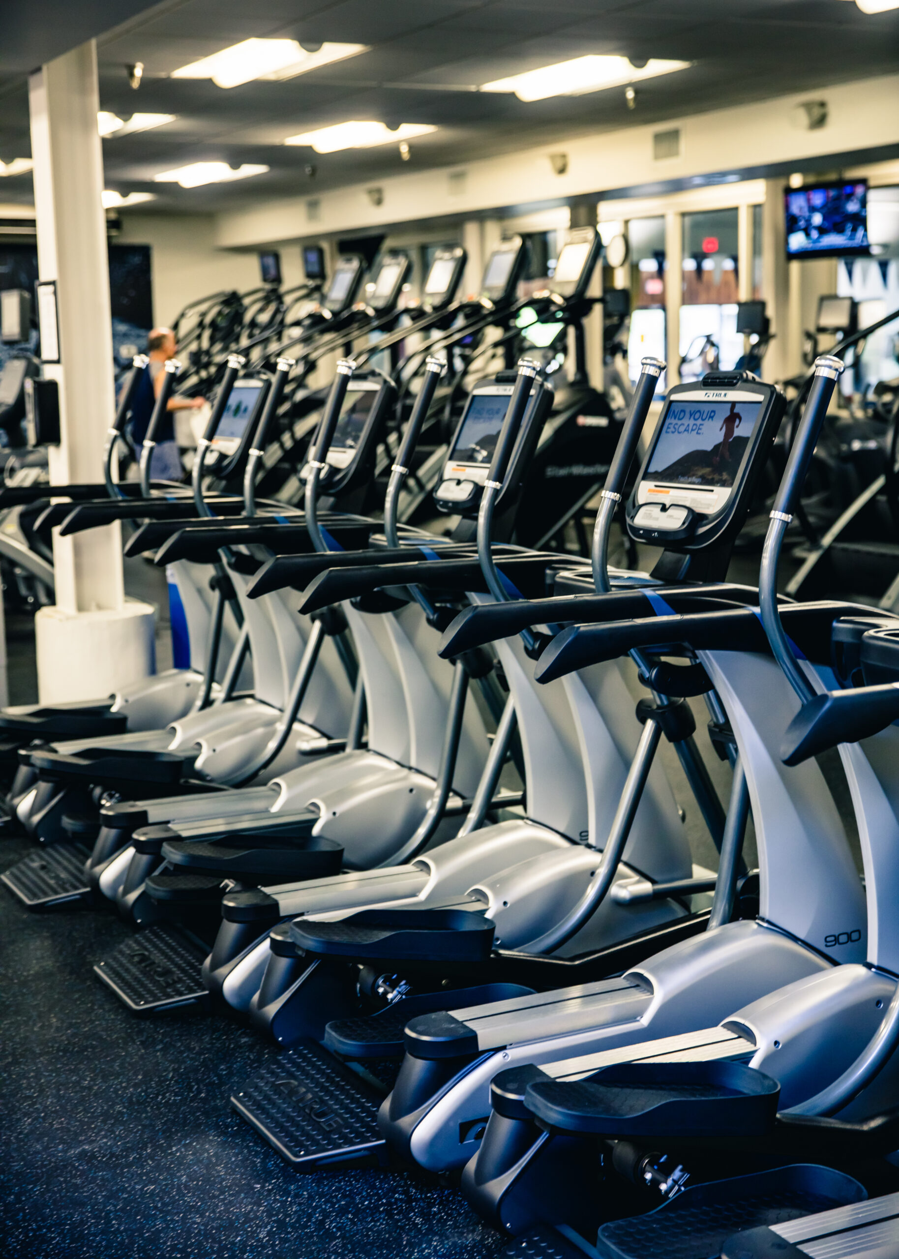 Rows of elliptical machines with digital screens are lined up in a well-lit gym, with additional fitness equipment and mirrors visible in the background.