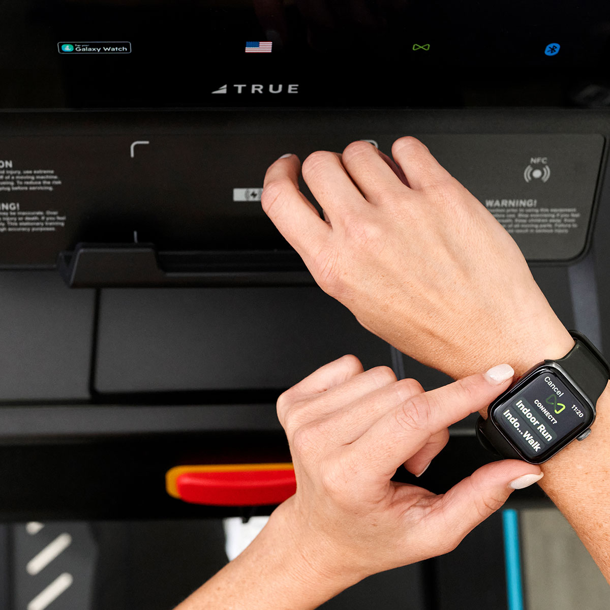 A person stands in front of a treadmill, holding their left wrist with their right hand and viewing fitness tracking information on a smartwatch. The treadmill display and smartwatch are both visible.