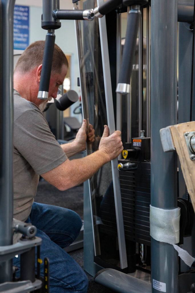 A man kneels and repairs a weight machine at a gym, holding a metal panel in place near the weight stack. Tools and other exercise equipment are visible in the background.