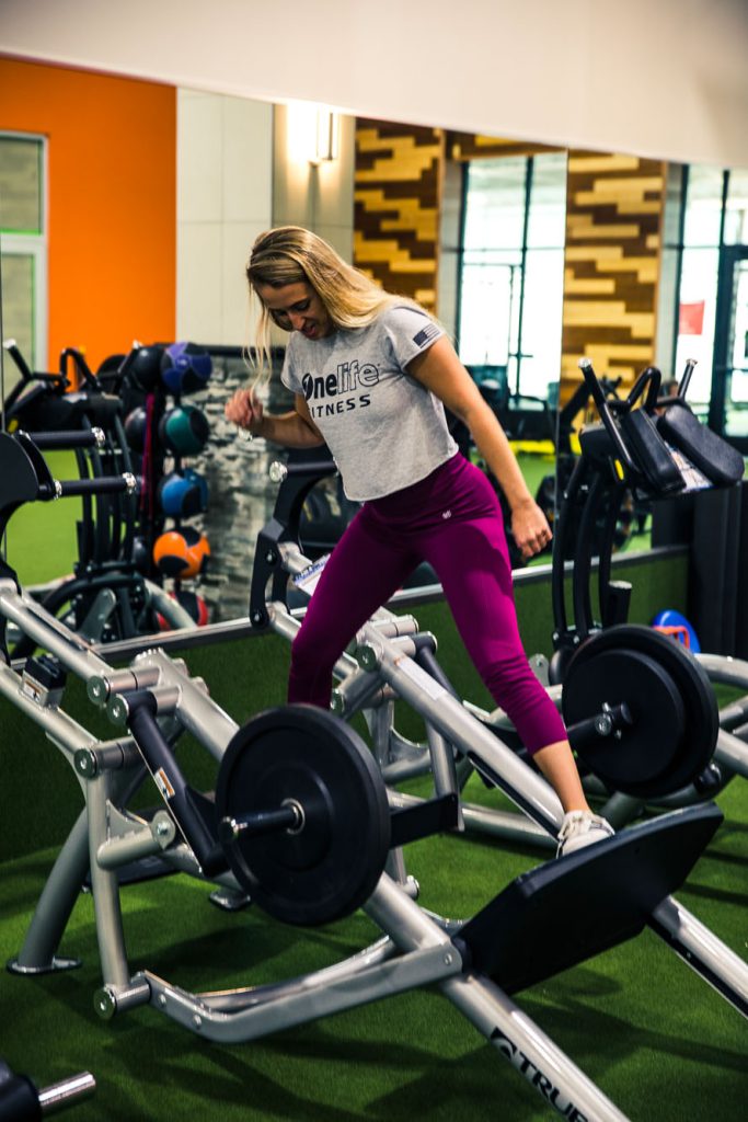 A woman in a gray Onelife Fitness shirt and purple leggings steps onto a leg press machine in a brightly lit gym with colorful walls and various fitness equipment.