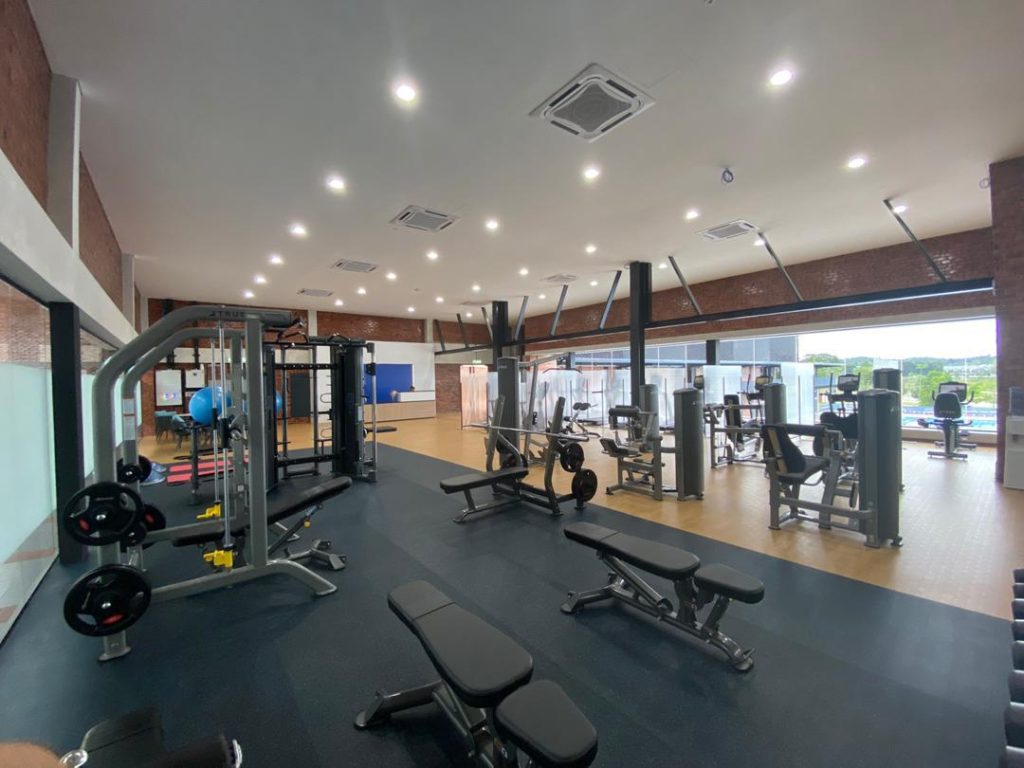 Spacious gym with various exercise machines, free weights, benches, and large windows letting in natural light. The room has a light wood floor, brick walls, and modern lighting throughout.