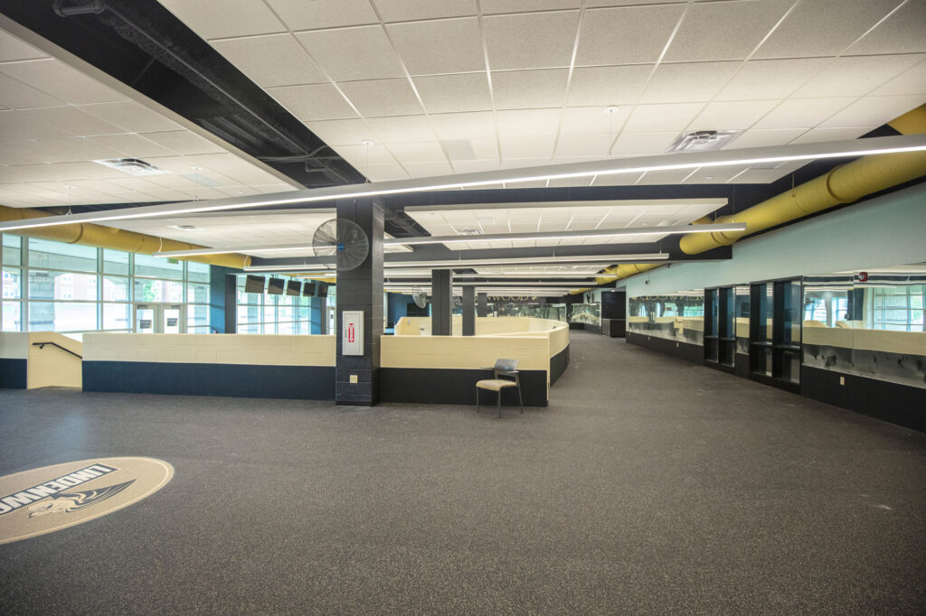 Large, modern indoor space with black and yellow accents, empty except for a single chair. Features wide windows, a circular logo on the floor, mirrors lining one wall, and exposed ceiling ducts.