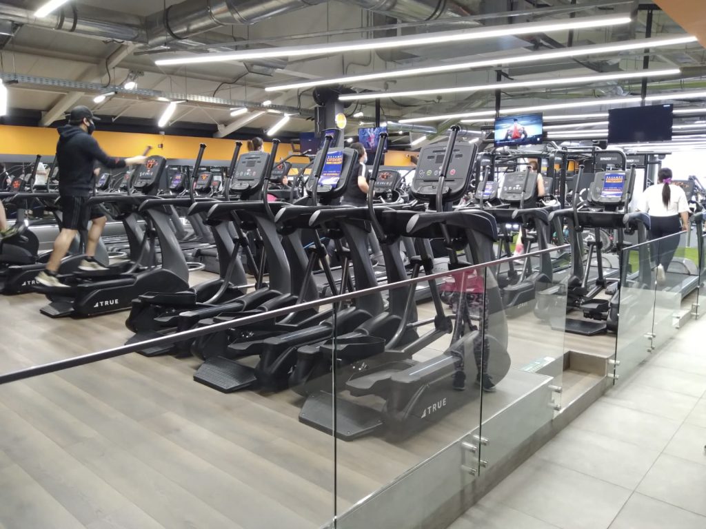 A modern gym with several people using treadmills and elliptical machines. The equipment faces large mirrors and TV screens, and the area is separated by a glass barrier. The gym has bright lights and an industrial ceiling.
