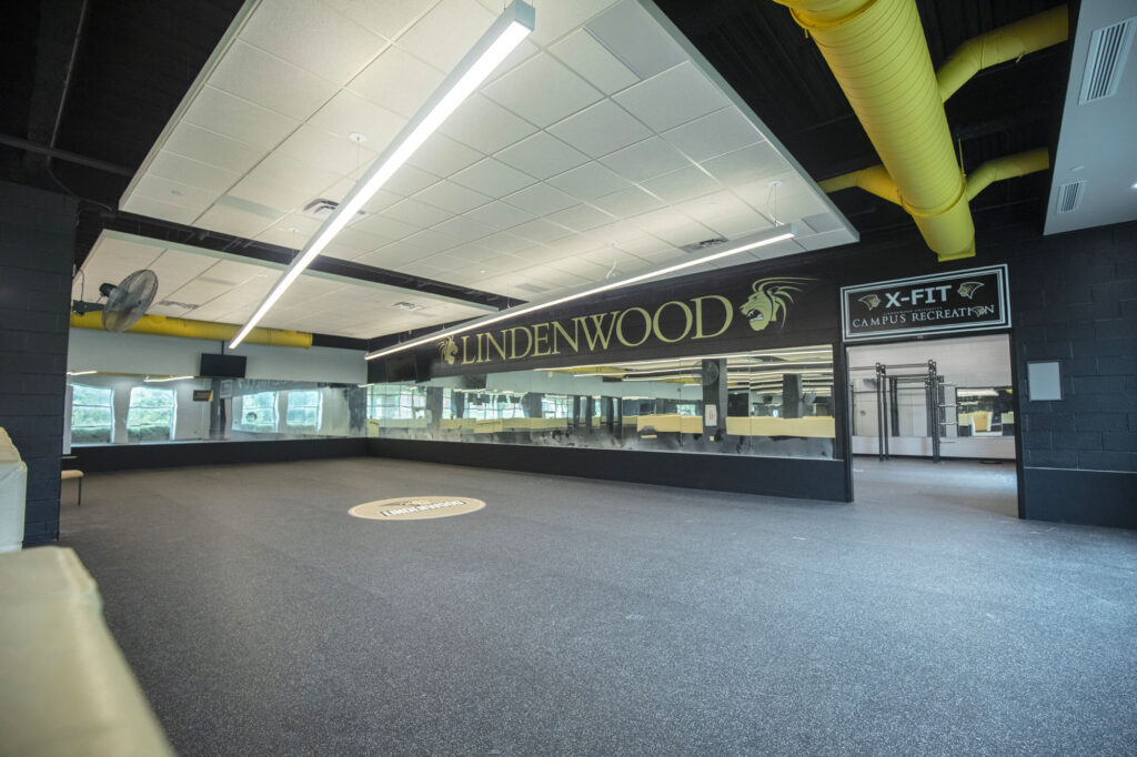 Spacious indoor gym area with black floors, yellow ceiling pipes, mirrored walls, and LINDENWOOD and X-FIT Campus Recreation signs. Large windows let in natural light. Room is empty.