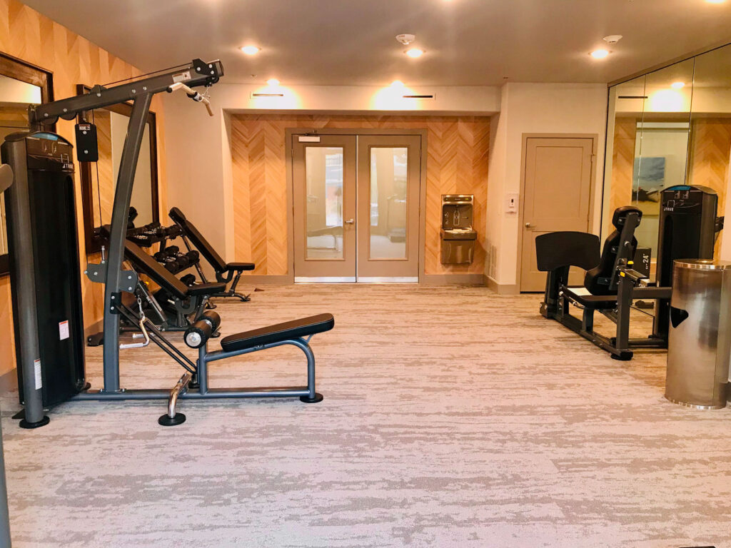 A small fitness room with exercise machines including a weight bench and leg press, beige carpeting, mirrored walls, double doors, and a water fountain at the back.