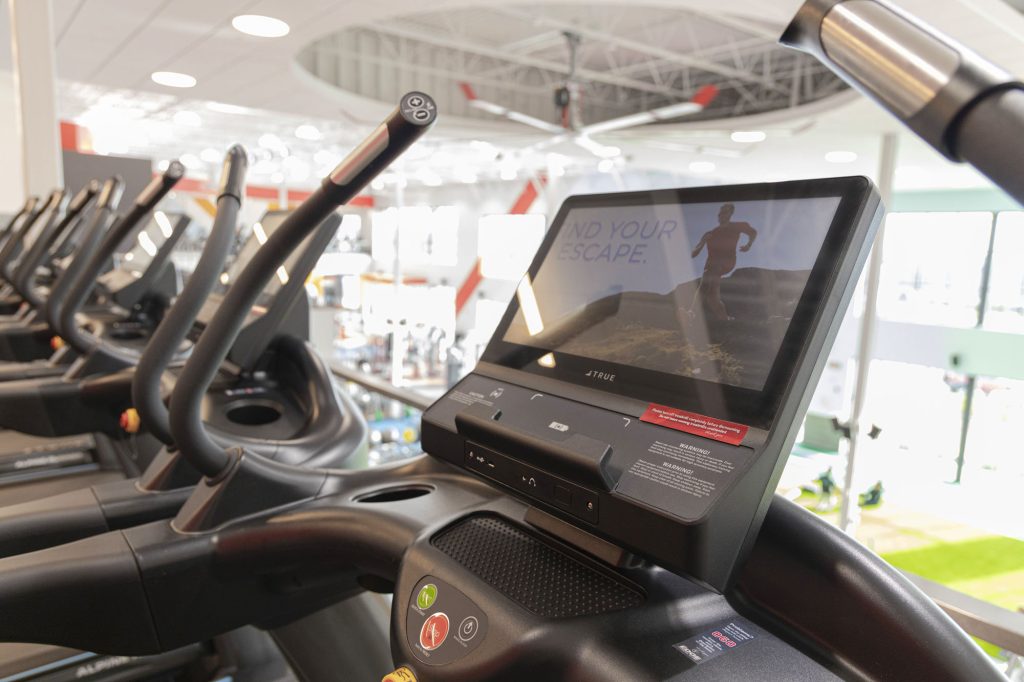 A close-up of a treadmill display screen showing a video of a person running outdoors. Several treadmills are lined up in a brightly lit gym with large windows in the background.