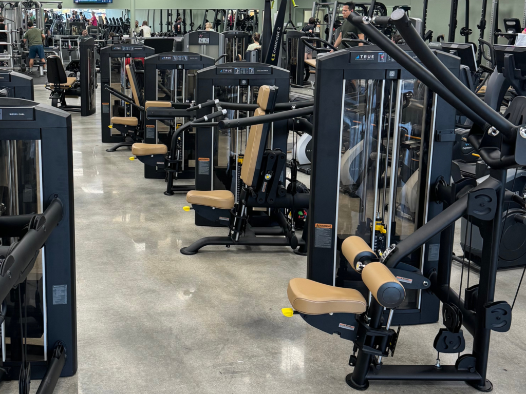 Rows of modern exercise machines with tan seats are arranged neatly in a gym with shiny floors. People can be seen working out in the background among the various fitness equipment.